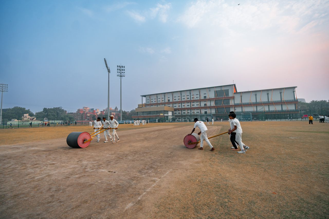 Groundskeepers preparing cricket field in Delhi, India with rollers at sunset.