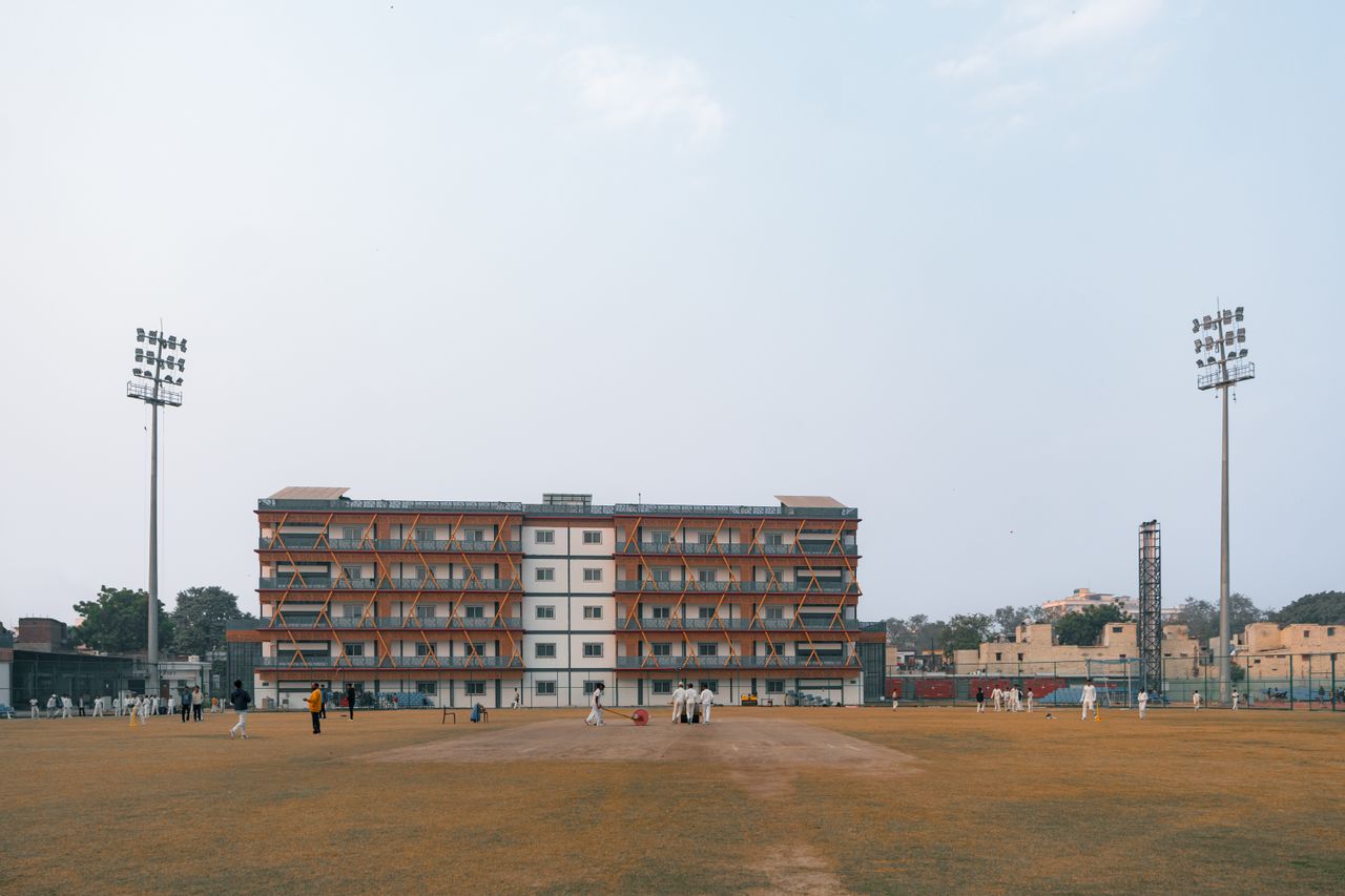 Cricket match in progress at a sports field in Delhi, India, with players and spectators under clear skies.