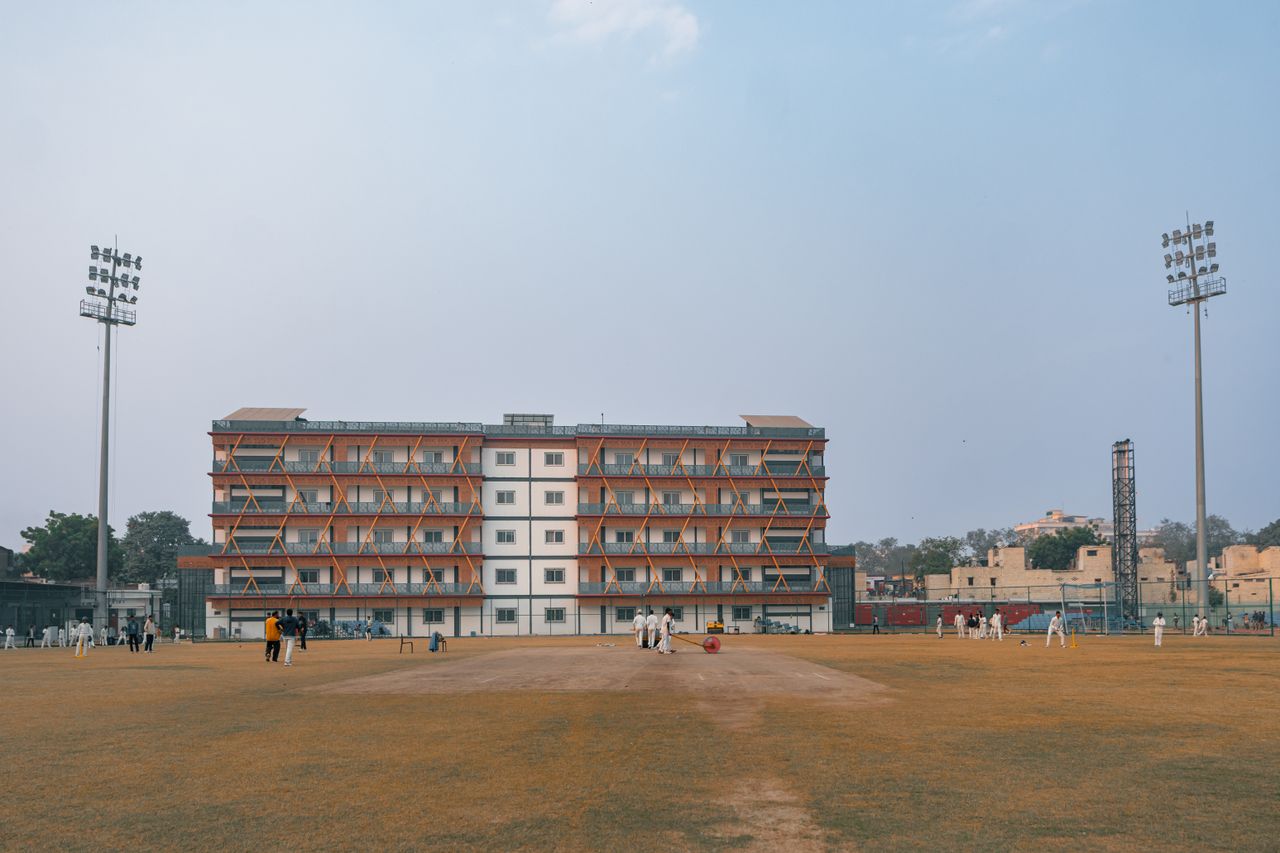 Daytime cricket match on a large sports field in Delhi, India with players and floodlights.