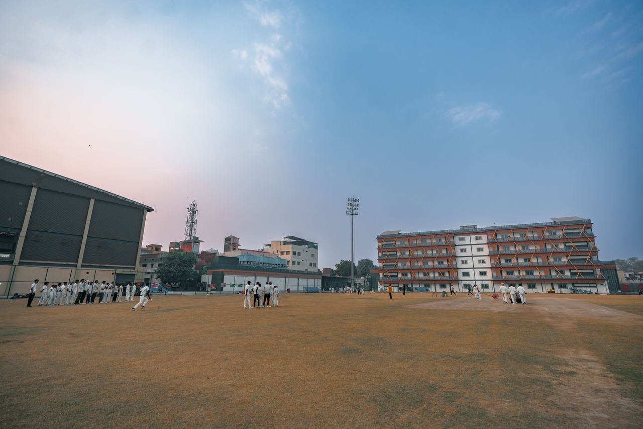 Outdoor cricket match on a sports field with players under a clear sky in Delhi, India.